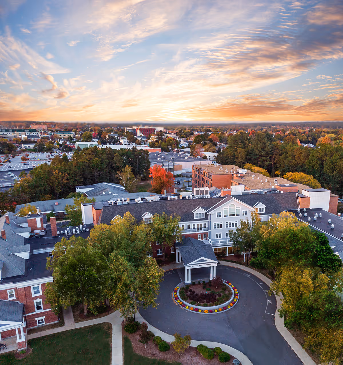 Aerial view of Hunt Community senior living facility at sunset, showing a large building with a circular driveway entrance surrounded by trees and landscaping, with a colorful sky and a suburban area in the background.