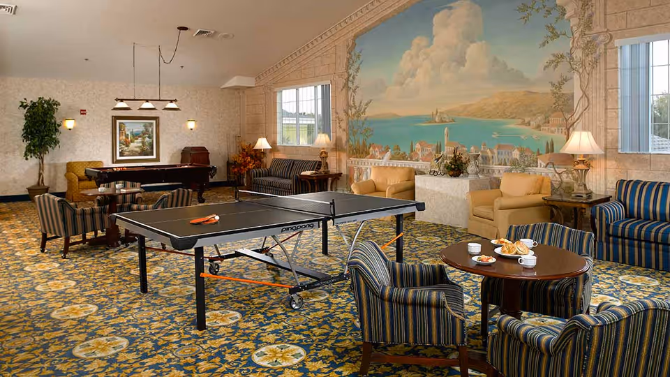 A spacious common room with a ping pong table in the center, surrounded by striped and solid armchairs and sofas. There is a round table with cups and plates of food, a pool table in the background, and a large mural on the wall depicting a coastal scene with buildings and water. The room has patterned carpet and several lamps providing warm lighting.
