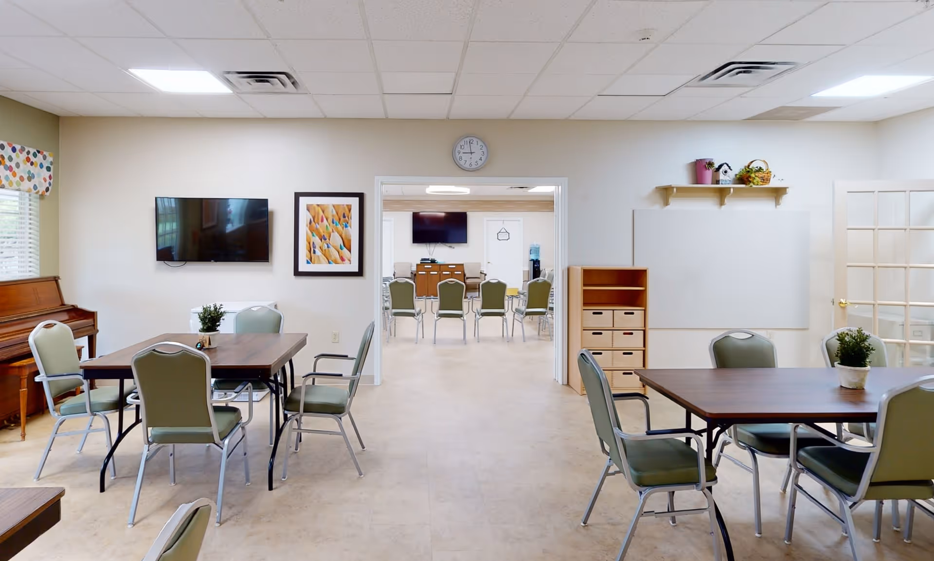 A bright, clean common area in a memory care facility featuring multiple tables with green cushioned chairs arranged around them. A piano is positioned against the left wall under a window with a colorful valance. A flat-screen TV and framed artwork hang on the wall. In the background, another room with more chairs and a TV is visible through a wide doorway. The room has a clock above the doorway, a small shelving unit, a whiteboard, and a shelf with decorative items on the right wall.