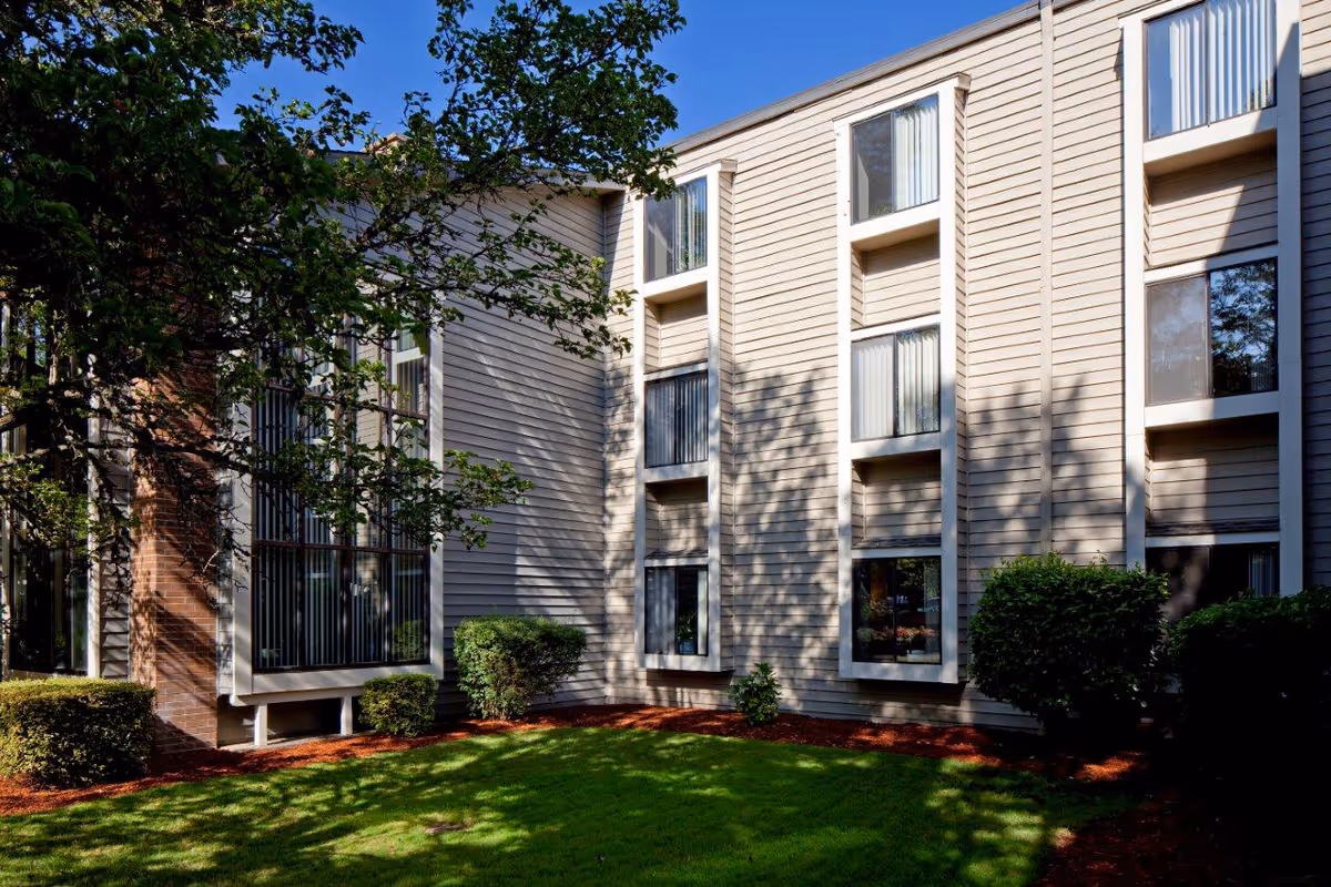 Exterior view of a multi-story residential building with beige siding, multiple windows, and a well-maintained lawn with bushes and trees casting shadows on the building.