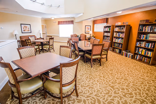 A well-lit common area with multiple wooden tables and chairs arranged on a patterned carpet. The room features bookshelves filled with books against an orange accent wall, framed artwork, and windows with brown blinds. The space appears cozy and inviting, suitable for reading or socializing.