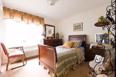 A cozy bedroom with a wooden bed frame, a striped and floral bedspread, and multiple pillows. There is a wooden dresser with decorative items and a framed picture on the wall above the bed. A window with a brown valance lets in natural light, and a wooden chair is placed near the window. A decorative metal shelf with framed photos and plants is visible on the right side of the room.