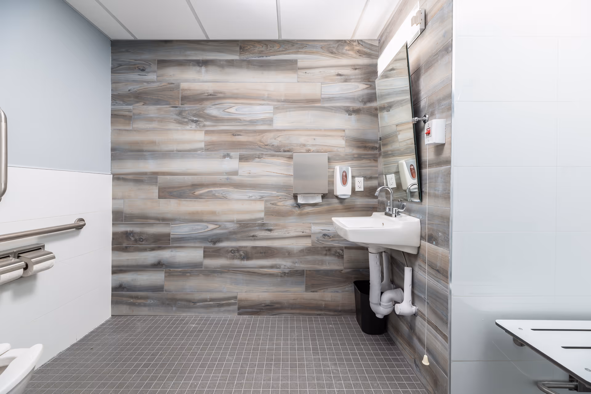 A clean, modern accessible bathroom with wood-patterned wall tiles, a white sink with exposed plumbing, a large mirror, a paper towel dispenser, soap dispenser, and a grab bar next to a toilet seat. The floor is covered with small square gray tiles.