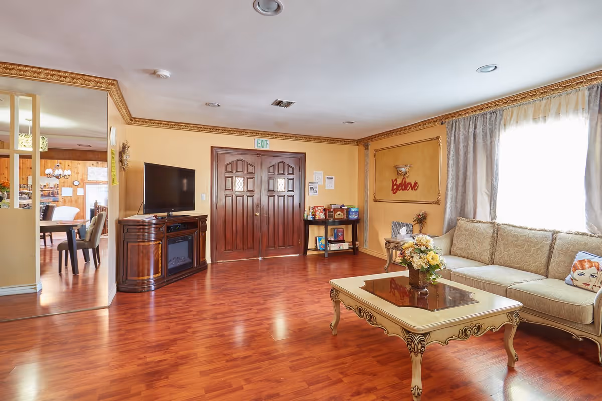A cozy senior living facility common area with polished wooden floors, a beige ornate sofa with patterned cushions, a decorative coffee table with a flower arrangement, a flat-screen TV on a wooden cabinet, double wooden doors with an exit sign above, and a small table with board games and books. The walls are painted yellow with gold trim, and a window with sheer curtains lets in natural light.