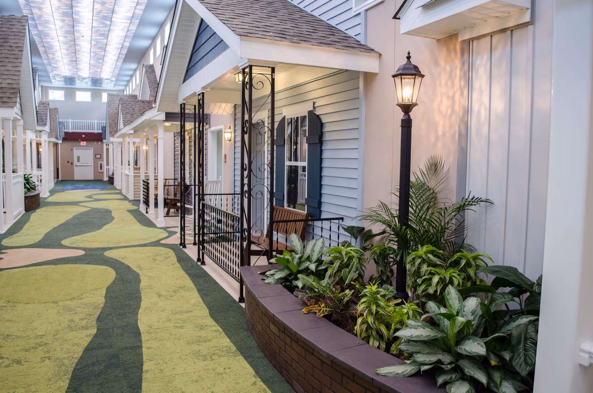 Indoor corridor styled like a streetscape with cottage-front doors, porch benches, plants, and a decorative lamppost.