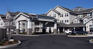 Exterior view of a large multi-story senior living facility building with a covered entrance and several parked cars in front under a clear blue sky.
