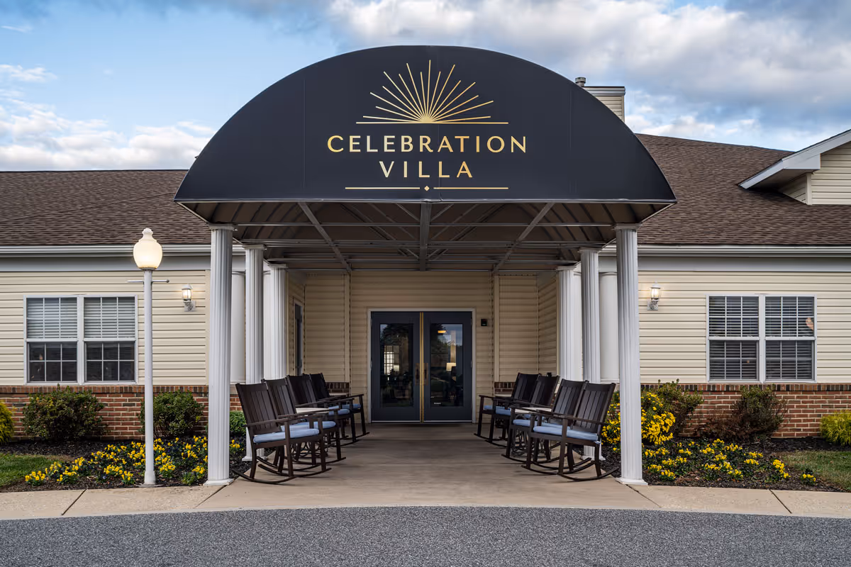 Front entrance of Celebration Villa featuring a covered black awning with the facility name, white columns and rocking chairs along the walkway.