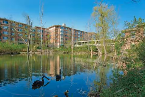 A pond with reflections, leafless trees, and a brick senior living campus in the background.