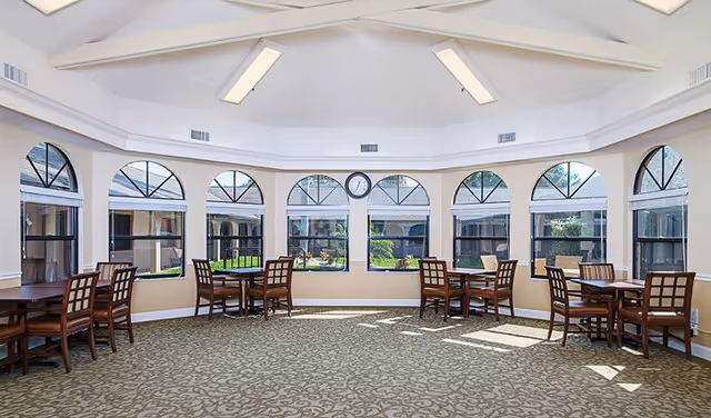 Sunlit circular dining area with multiple wooden tables and chairs arranged before arched windows overlooking a courtyard.