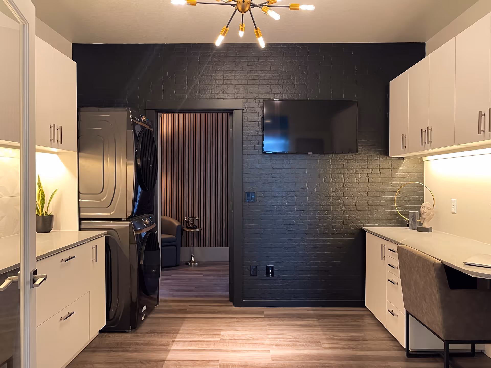 Modern interior room with stacked washer and dryer, white cabinets and countertops, a desk chair, and a wall-mounted TV on a dark brick wall.