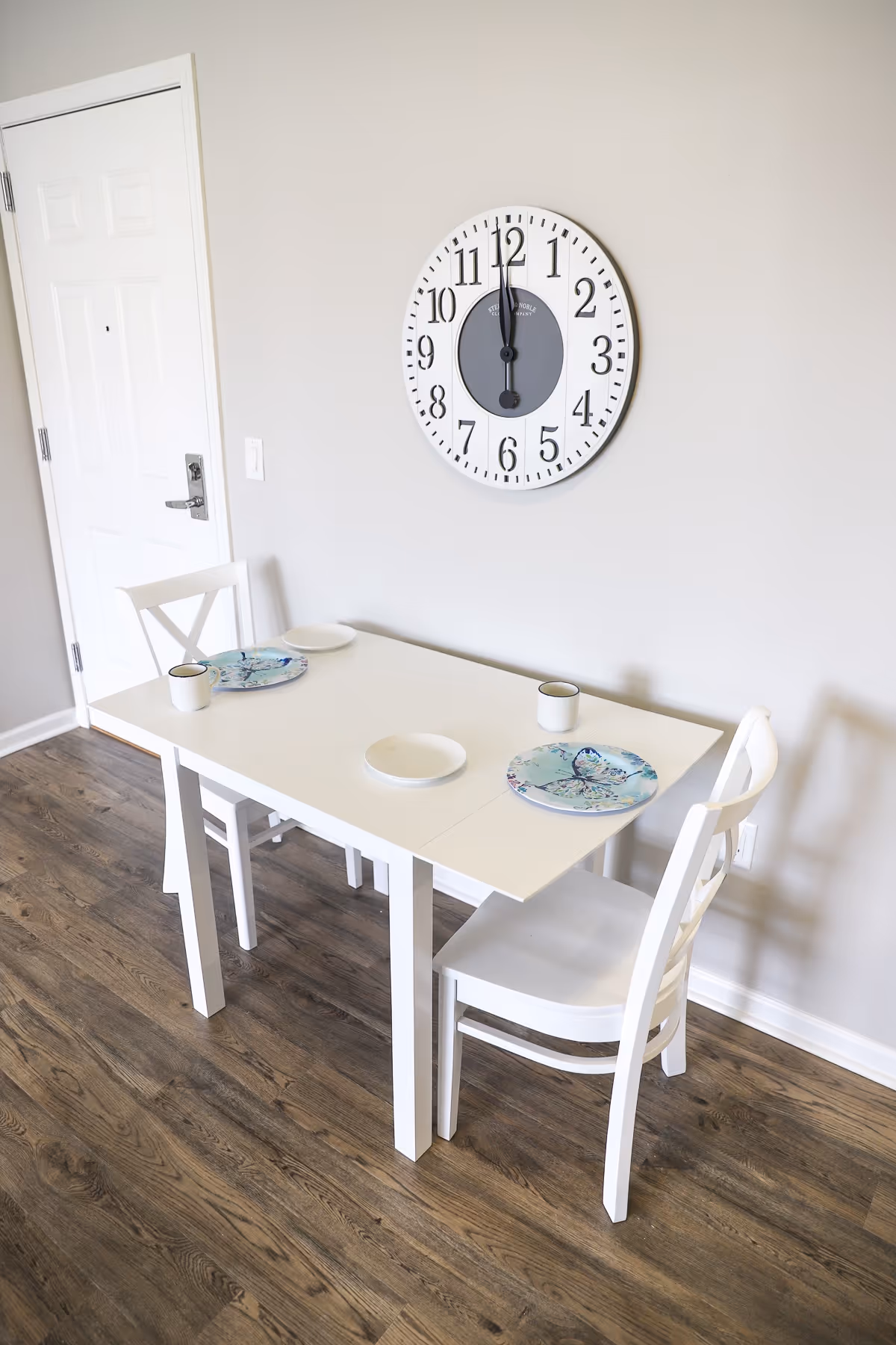 A small dining area with a white table set for two, featuring two white chairs, two plates with butterfly designs, two white mugs, and two empty white plates. A large round wall clock showing 12:00 is mounted on a light gray wall above the table. The floor is wooden, and a white door is visible in the background.