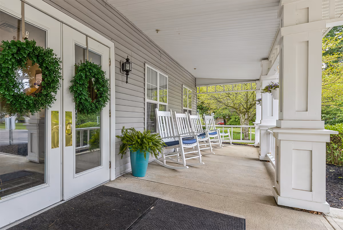 Covered porch area with white double doors decorated with green wreaths, several white rocking chairs with blue cushions lined up along the wall, a blue planter with a green fern, and hanging flower pots. Trees and greenery are visible in the background.