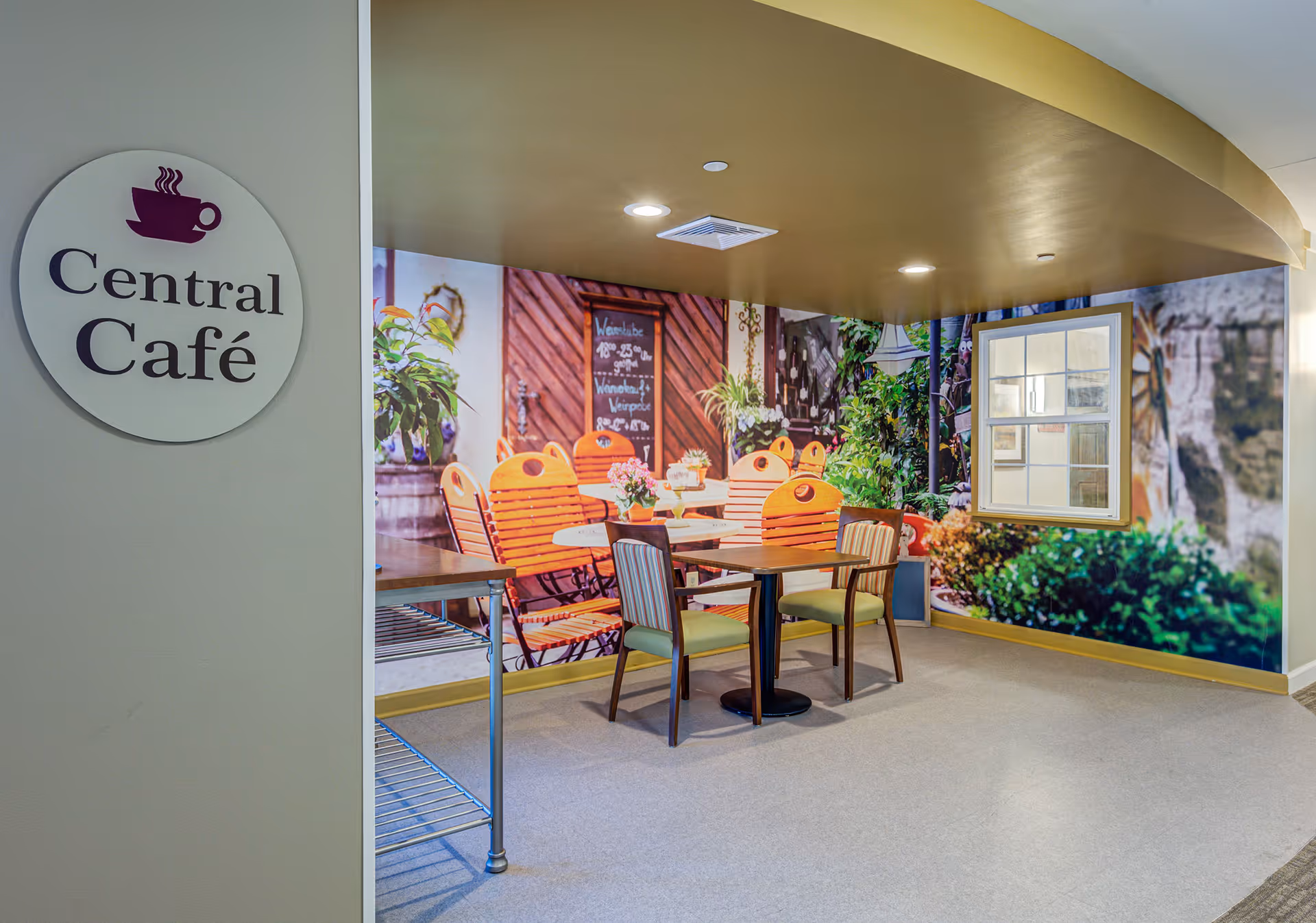 Indoor seating area labeled Central Café with a table and four chairs. The wall behind the seating area features a large mural depicting an outdoor café scene with orange chairs, plants, and a window.