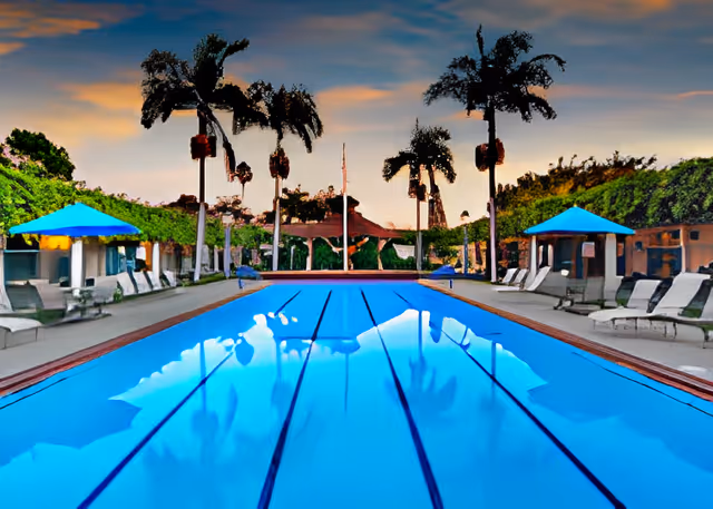 Outdoor swimming pool at sunset with clear blue water, surrounded by lounge chairs and umbrellas, tall palm trees in the background, and a gazebo at the far end.