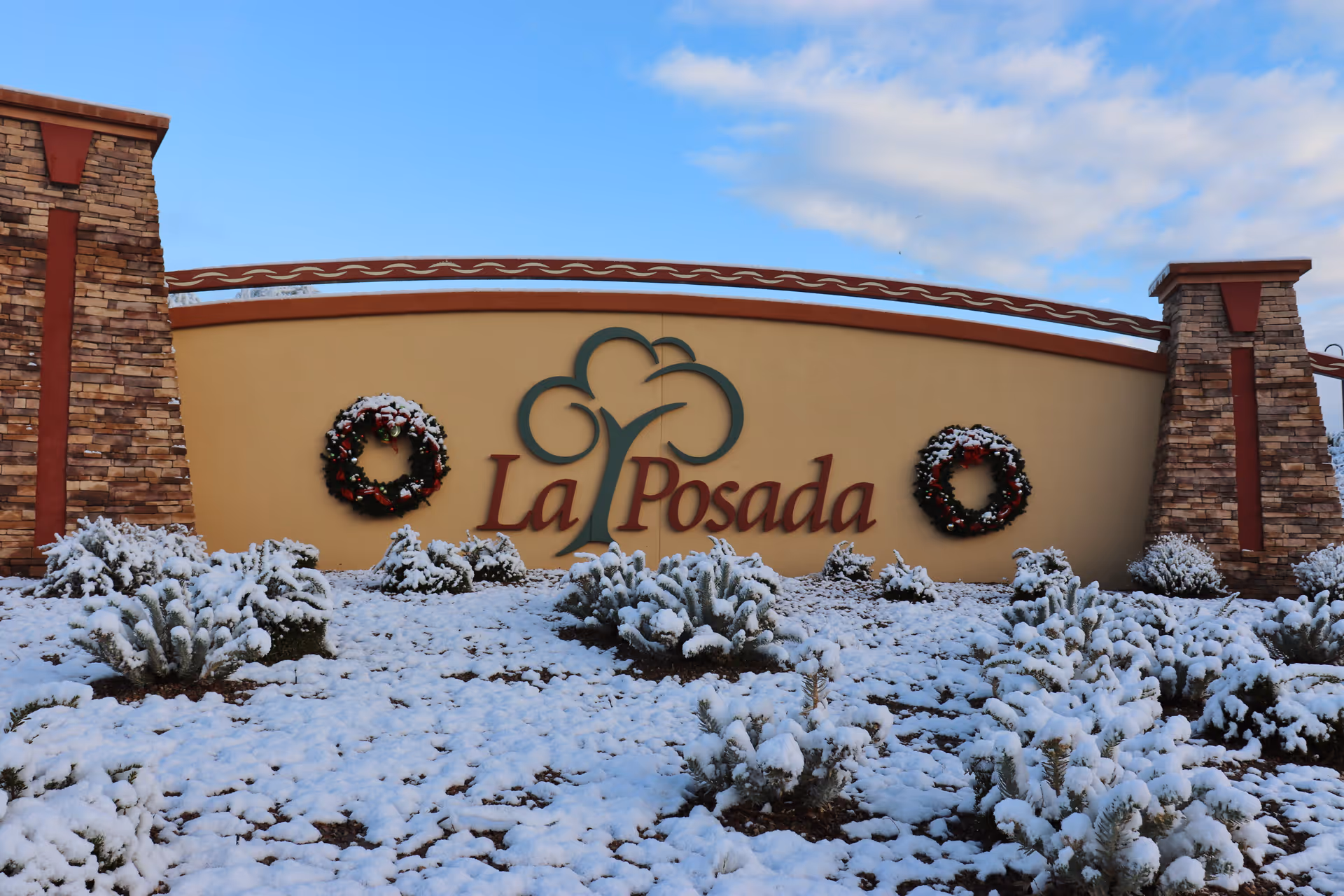 Entrance sign for La Posada at Green Valley with two stone pillars on each side, decorated with two Christmas wreaths, and snow-covered bushes in front under a partly cloudy sky.
