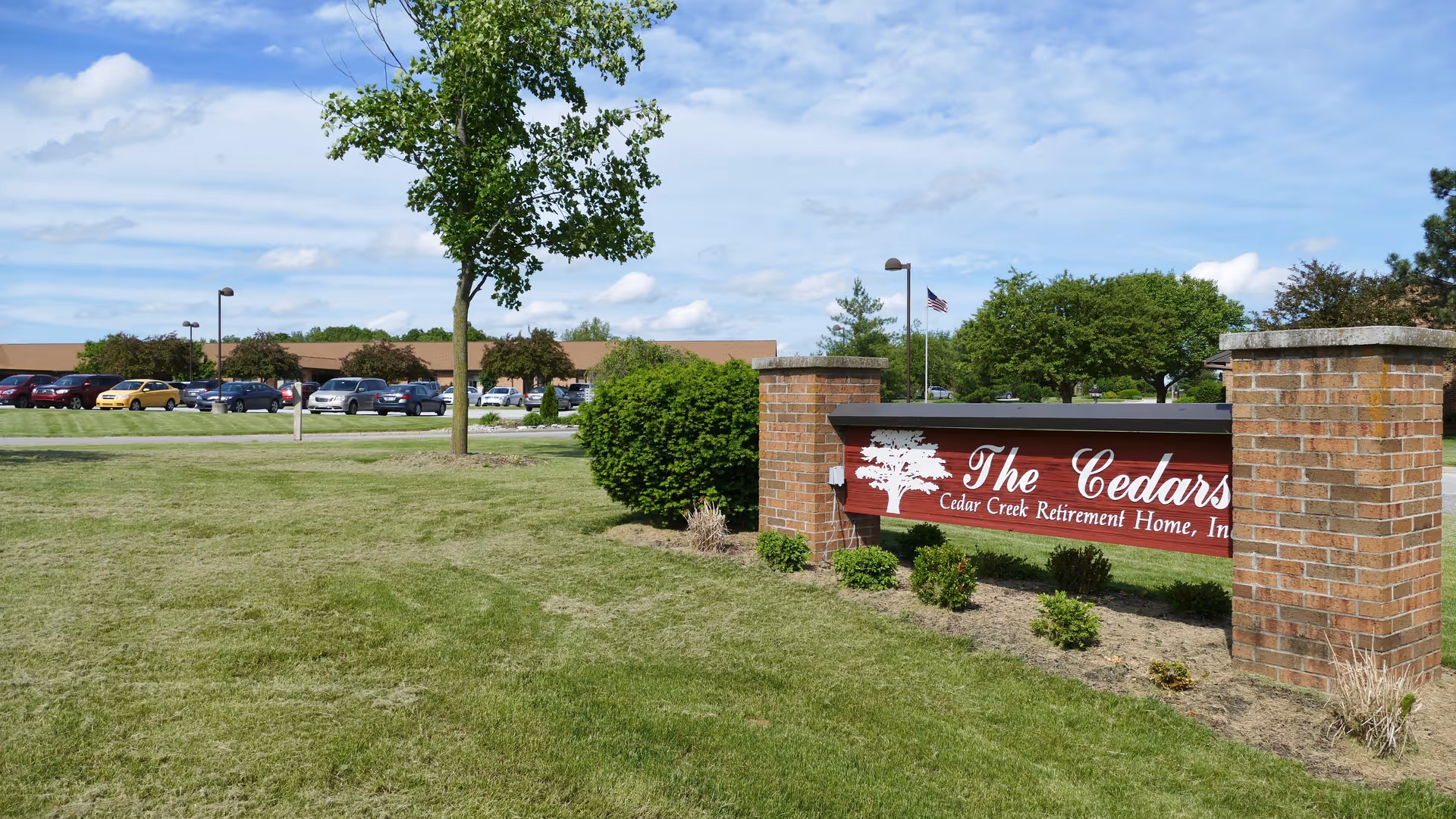 A brick sign reading 'The Cedars' sits on a grassy lawn with trees, a parking lot, and the facility building visible in the background.