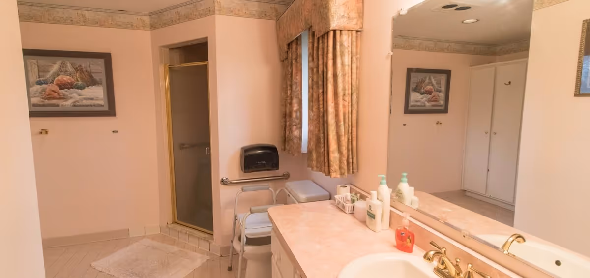 A bathroom with a shower stall with a glass door, a white toilet with a support frame, a countertop with a sink and various bottles of soap and lotion, a large mirror above the sink, a window with patterned curtains, a framed picture on the wall, and a white rug on the tiled floor.