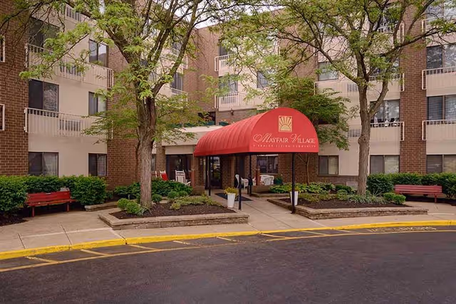 Front entrance of a brick senior living building with a red awning reading 'Mayfair Village', trees, benches, and planted beds.