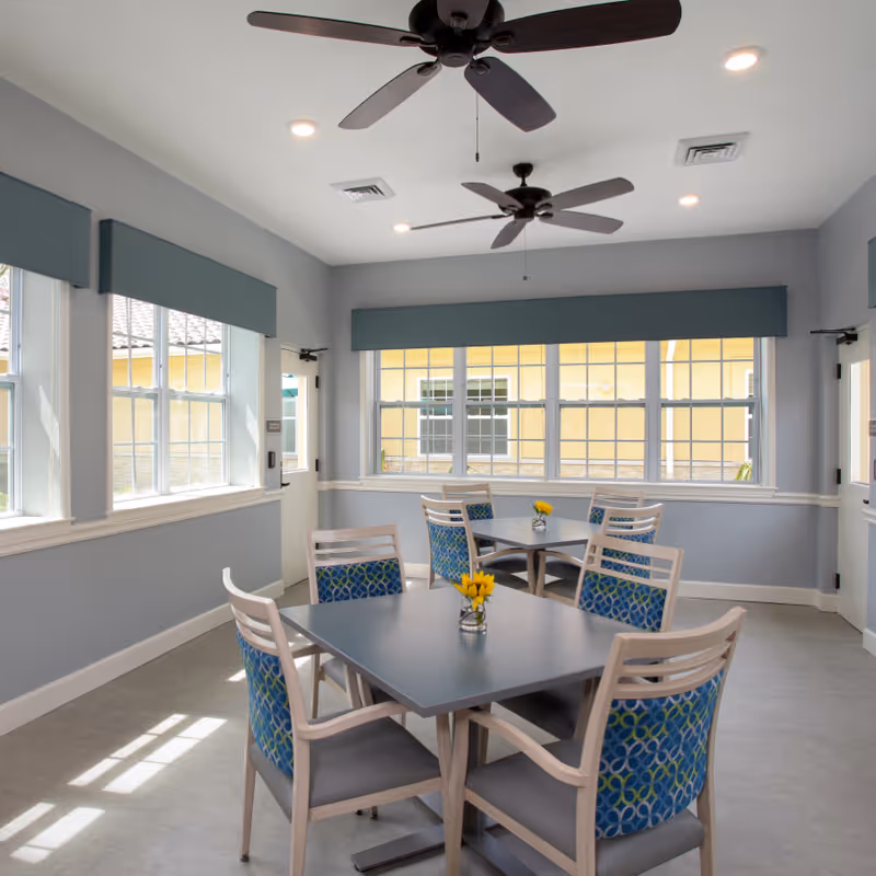 A bright dining room with two square tables, each surrounded by four chairs with blue patterned backs and gray seats. Small vases with yellow flowers are placed on each table. The room has large windows with blue valances, gray walls, and two ceiling fans with dark blades. Sunlight streams through the windows, casting shadows on the light-colored floor.