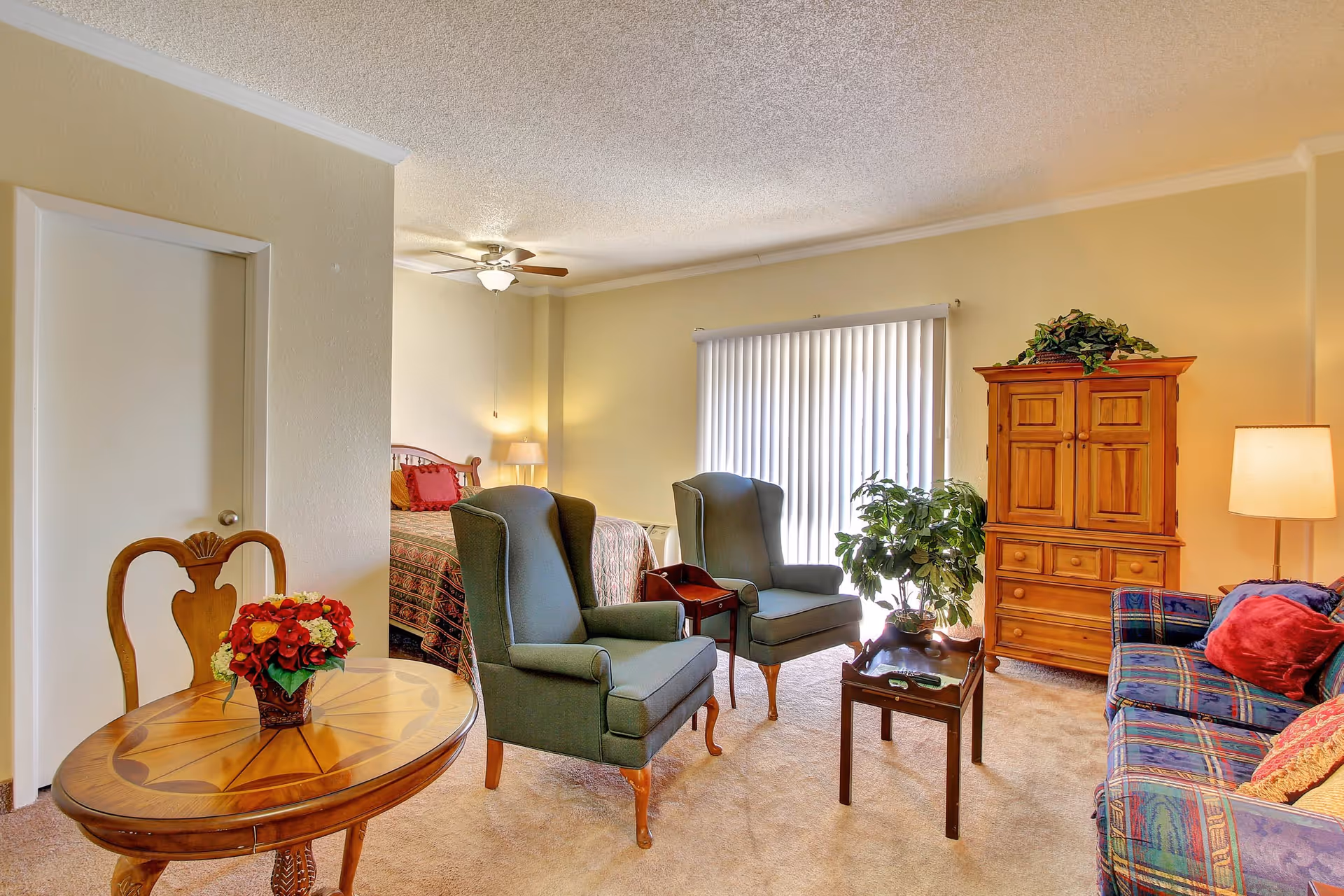 Bright senior living room with two green armchairs, a patterned sofa, wooden armoire, small tables including one with a flower arrangement, and a bed visible in the background.