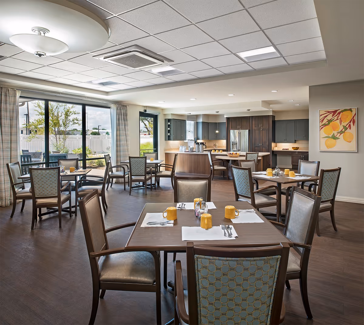 Bright communal dining room with tables and chairs set for meals and an open kitchen in the background next to large windows.