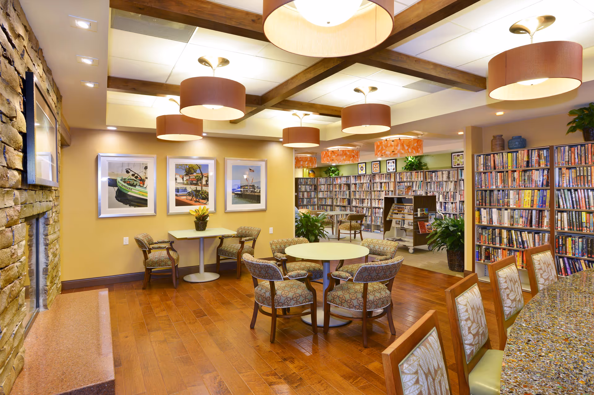 A cozy and well-lit common area in a retirement community featuring round tables with upholstered chairs, a stone fireplace on the left, framed pictures on the wall, and a large bookshelf filled with books in the background. The ceiling has wooden beams and multiple pendant lights with brown lampshades.