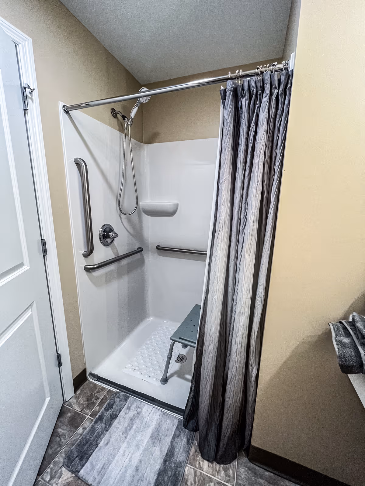 A small shower area with beige walls, a white shower base, and a gray shower curtain. The shower has a handheld showerhead, two horizontal grab bars, and a vertical grab bar. There is a small gray shower seat inside. A gray and white bath mat is placed on the tiled floor outside the shower.