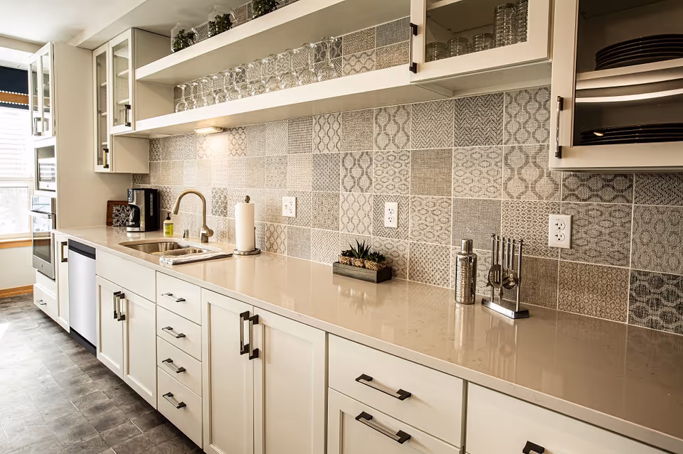 A modern kitchen with white cabinets, a beige countertop, and a patterned tile backsplash. The kitchen features a stainless steel sink with a faucet, a coffee maker, a paper towel holder, and various kitchen utensils. There are glass shelves with glasses and small potted plants above the countertop. Natural light is coming through a window on the left side.