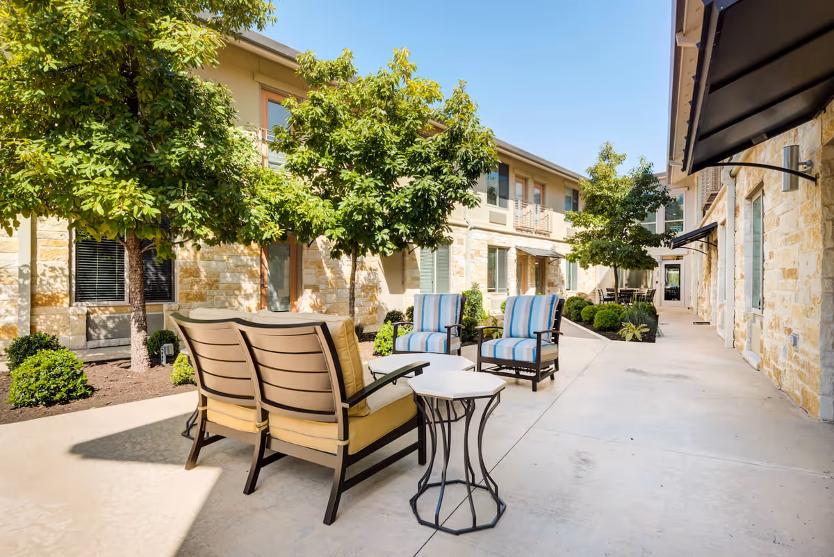 Outdoor courtyard with lounge chairs, small tables, and trees between stone-clad building facades.
