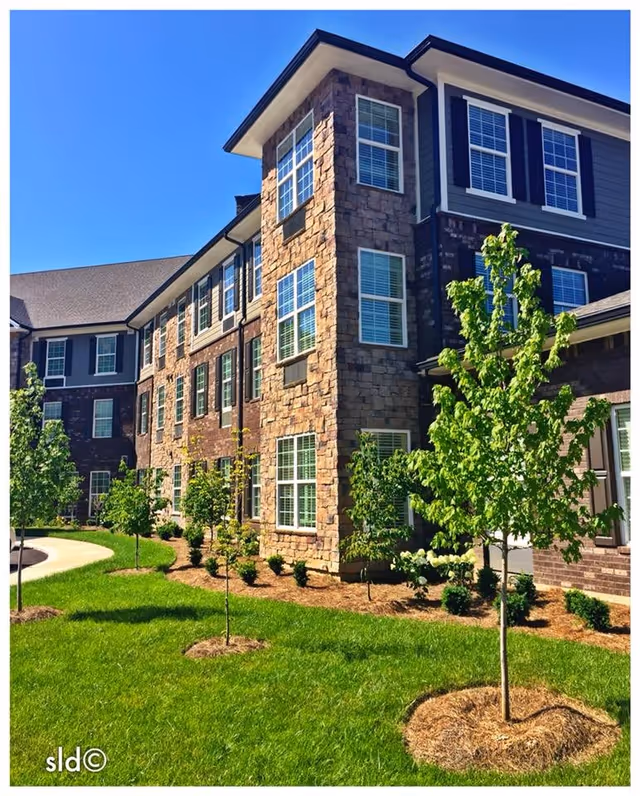Three-story stone-and-brick building exterior with many windows, landscaped lawn and young trees under a clear blue sky.