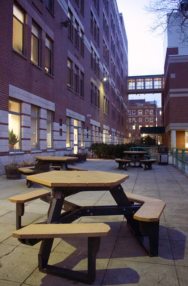 Outdoor courtyard between brick buildings with hexagonal picnic tables on a paved patio at dusk.