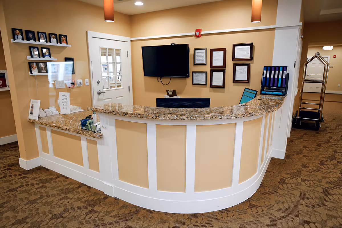 Reception desk area inside a senior living facility with a curved granite countertop, beige walls, and brown patterned carpet. Behind the desk is a wall-mounted TV, framed certificates, and a computer monitor. To the left, there are shelves with framed photos and a sign that reads 'PLEASE DO NOT LEAN ON PLEXI GLASS'. A luggage cart is visible in the hallway to the right.