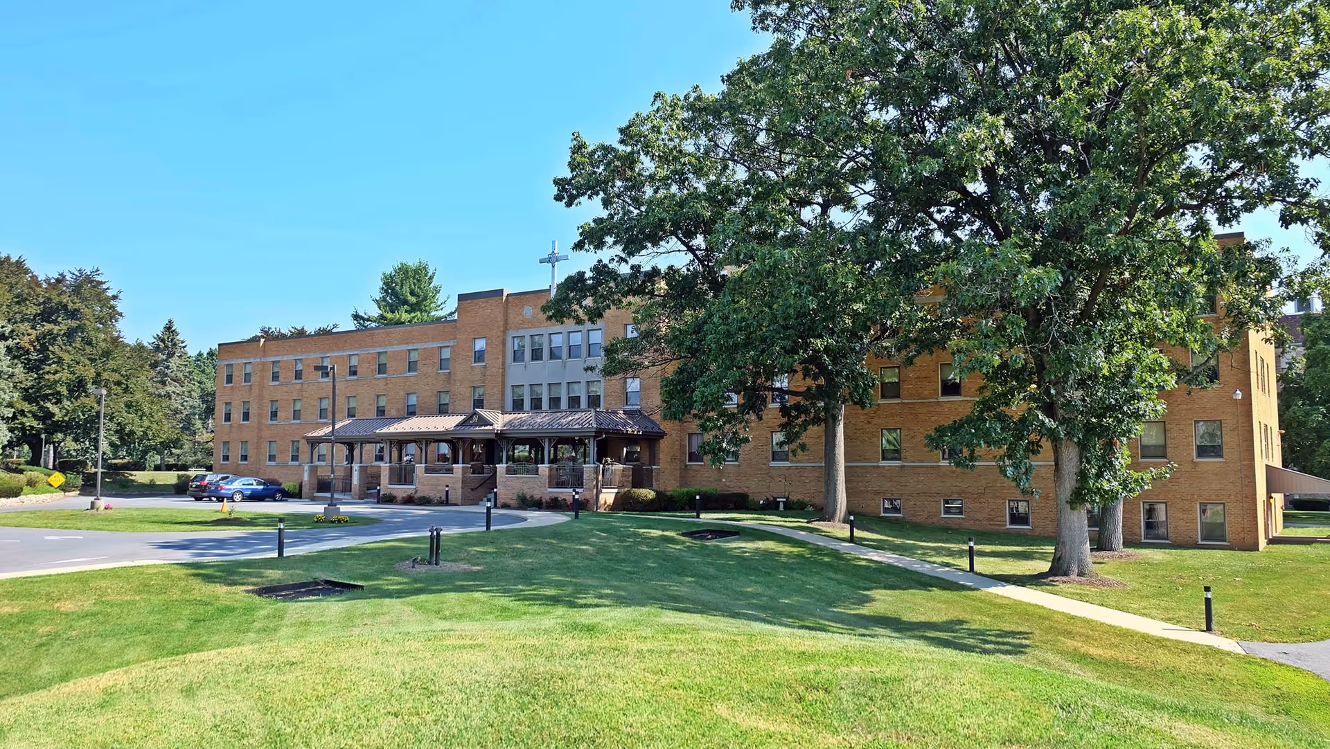 Exterior view of a three-story brick building with a covered entrance and a cross on the roof, surrounded by green grass, trees, and a parking area with several cars under a clear blue sky.