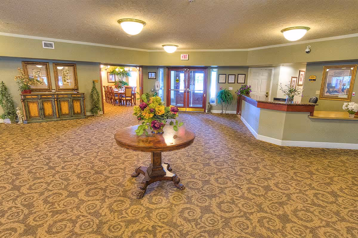 Spacious lobby area with patterned carpet, a round wooden table with a floral arrangement in the center, a reception desk to the right, and a dining area visible through an open doorway on the left. The entrance doors are straight ahead with exit signs above them. The walls are decorated with framed pictures and plants.