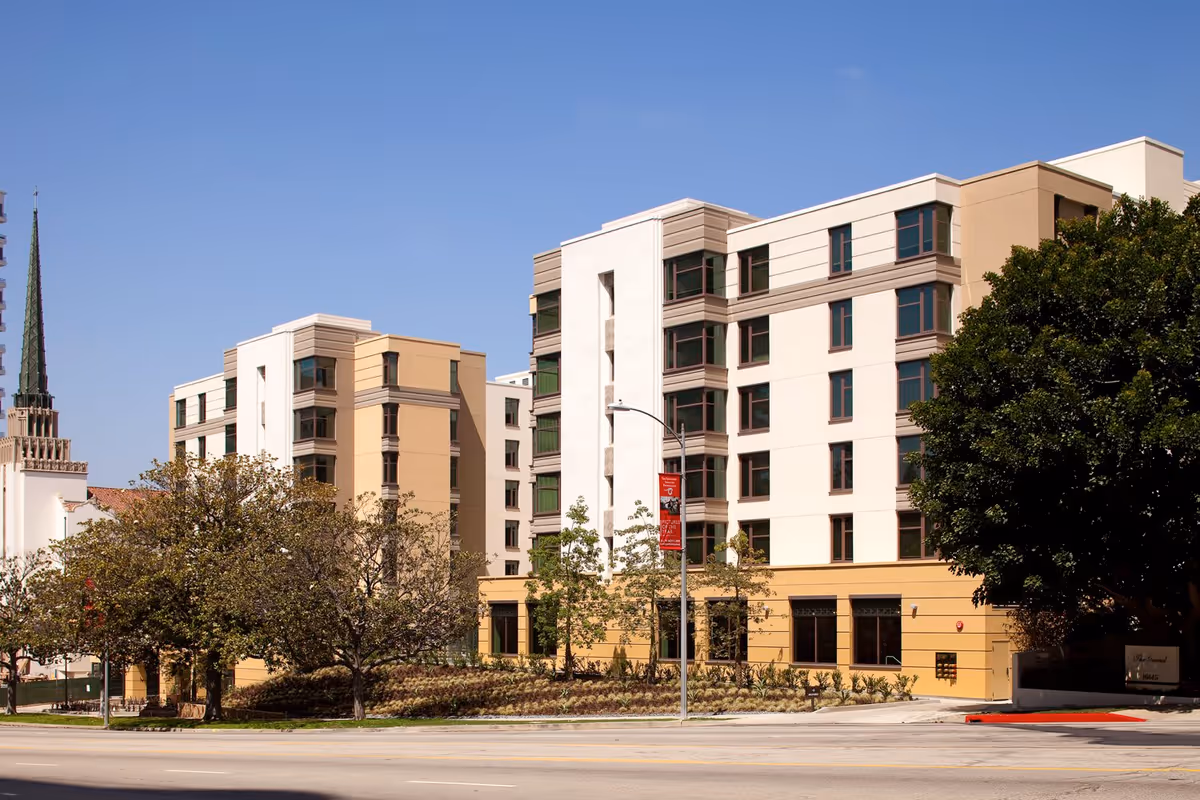 Exterior view of a multi-story senior living facility building with beige and white walls, surrounded by trees and landscaping under a clear blue sky.