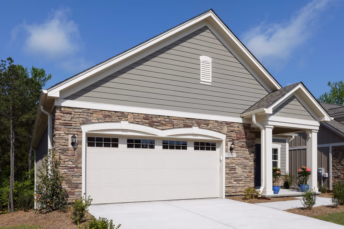 Exterior view of a single-story residential building with a two-car garage door, stone and siding facade, a small covered porch with white columns, and some landscaping including small bushes and potted plants.