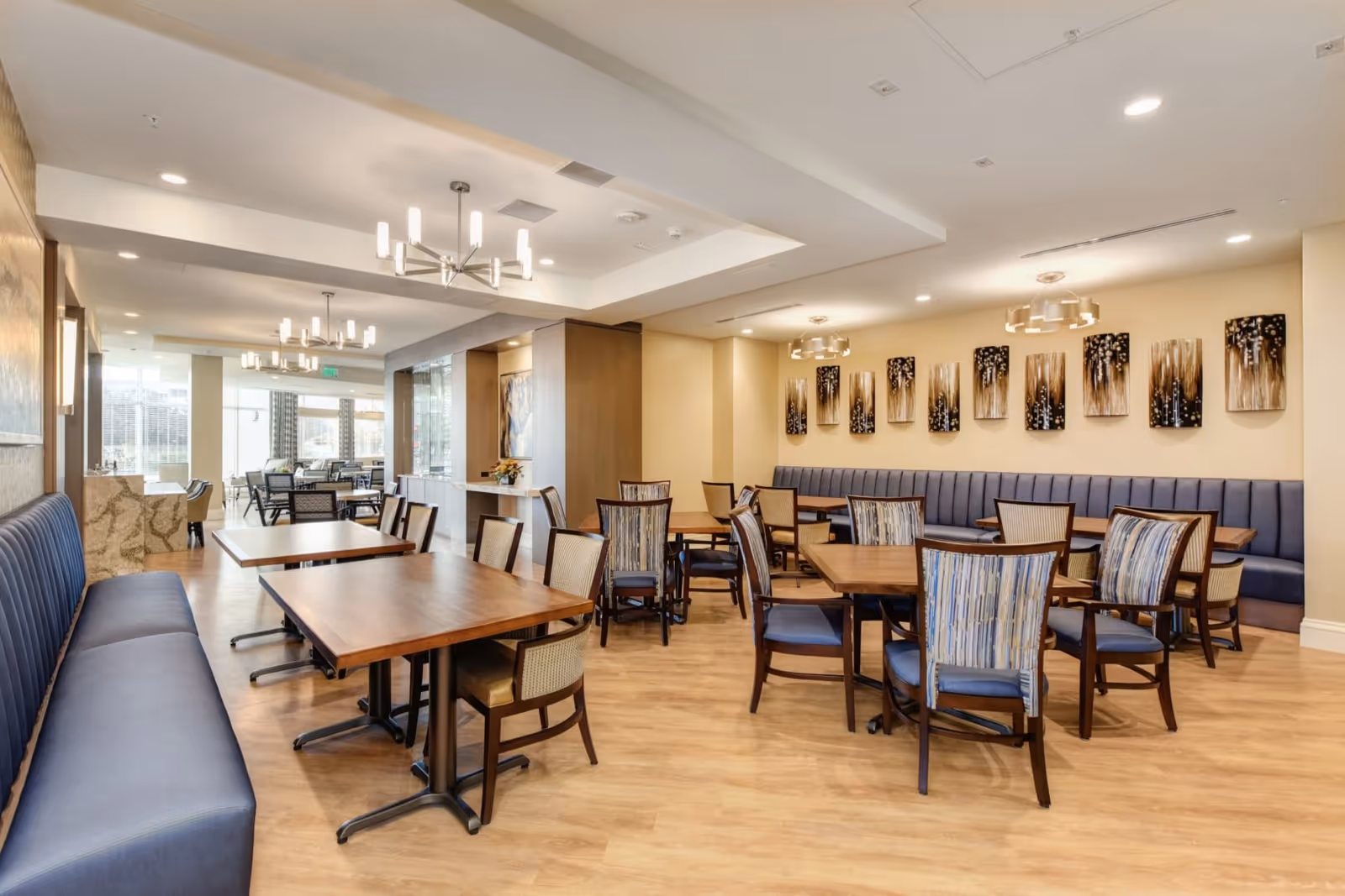 A spacious and well-lit dining room with wooden tables and a mix of blue cushioned chairs and banquette seating along the walls. Modern chandeliers hang from the ceiling, and decorative artwork is displayed on the walls. The room has light-colored walls and wood flooring, with large windows allowing natural light to enter.