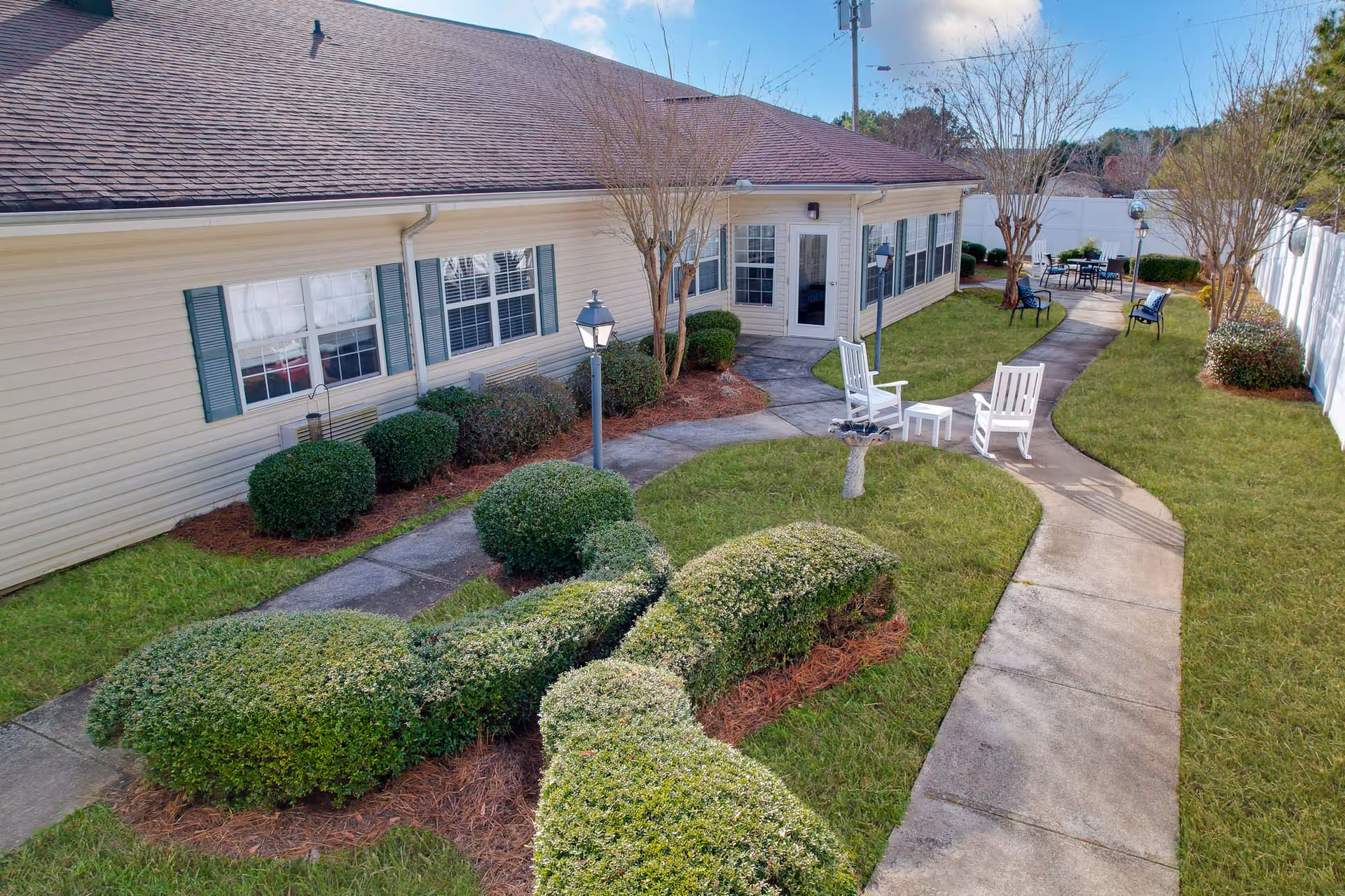 Outdoor courtyard area at PruittPlace - Wesleyan featuring a winding concrete pathway, neatly trimmed bushes, green grass, white rocking chairs with a small table, and a patio table with chairs in the background. The building has beige siding, green shutters, and multiple windows. There are also lamp posts and a white privacy fence surrounding the area.