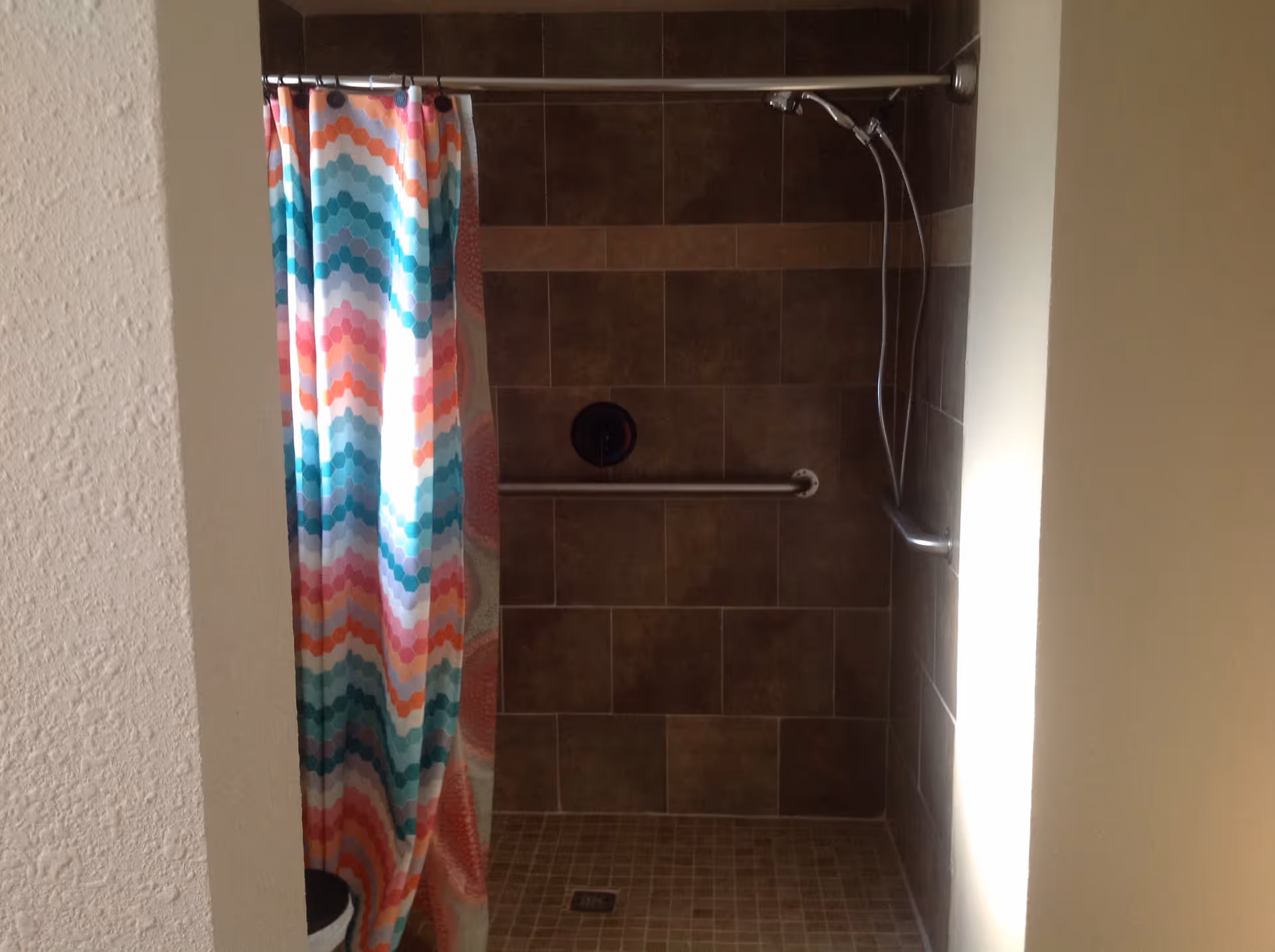 A tiled shower area with brown tiles on the walls and floor, featuring a silver grab bar and a handheld showerhead. A colorful shower curtain with a hexagonal pattern in shades of teal, orange, pink, and white is partially drawn to the left.