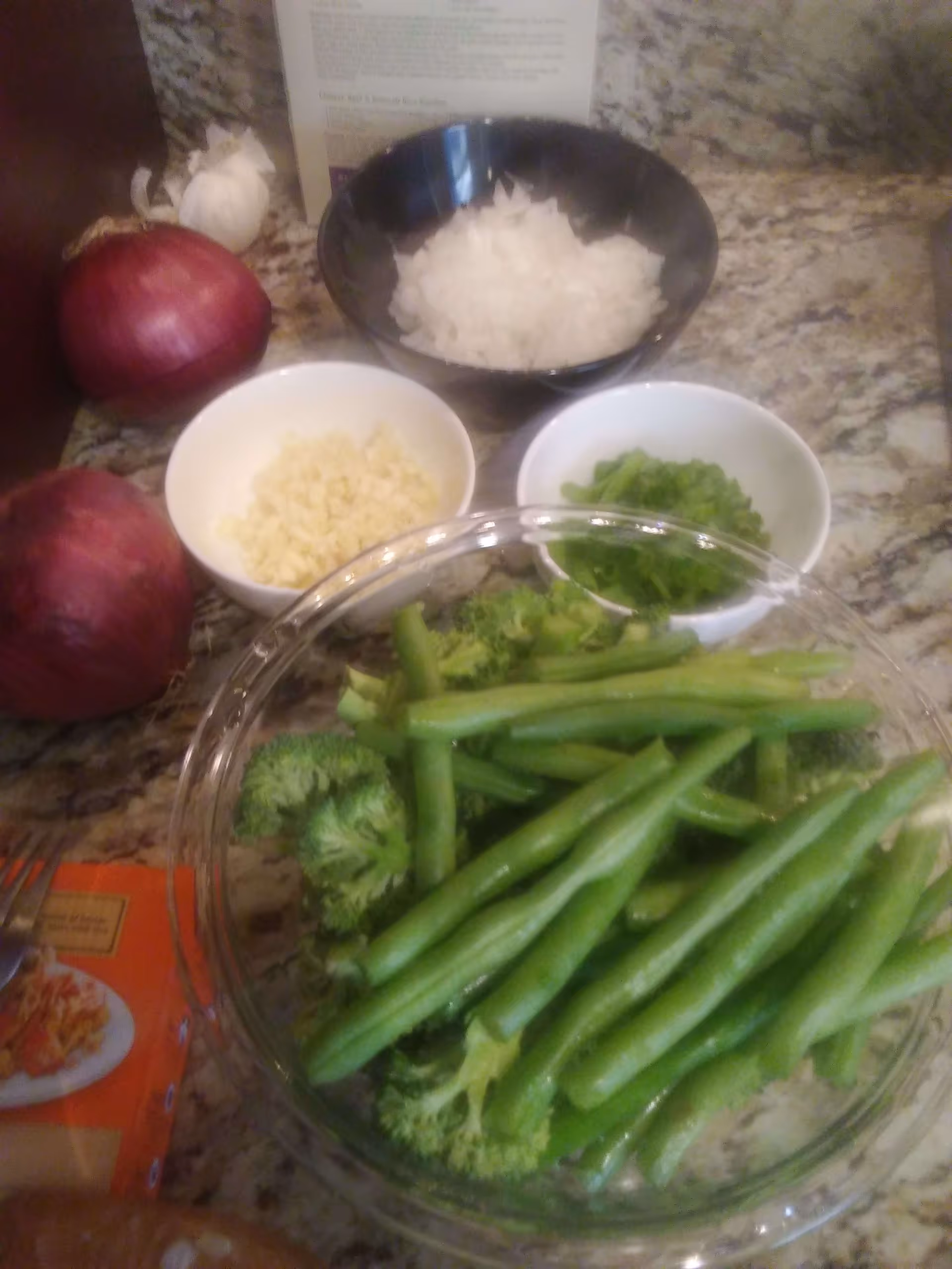 A kitchen countertop with various fresh vegetables and ingredients including two whole red onions, a bowl of chopped onions, a bowl of chopped garlic, a bowl of chopped green onions, and a glass bowl filled with broccoli and green beans. A recipe card is partially visible in the background.