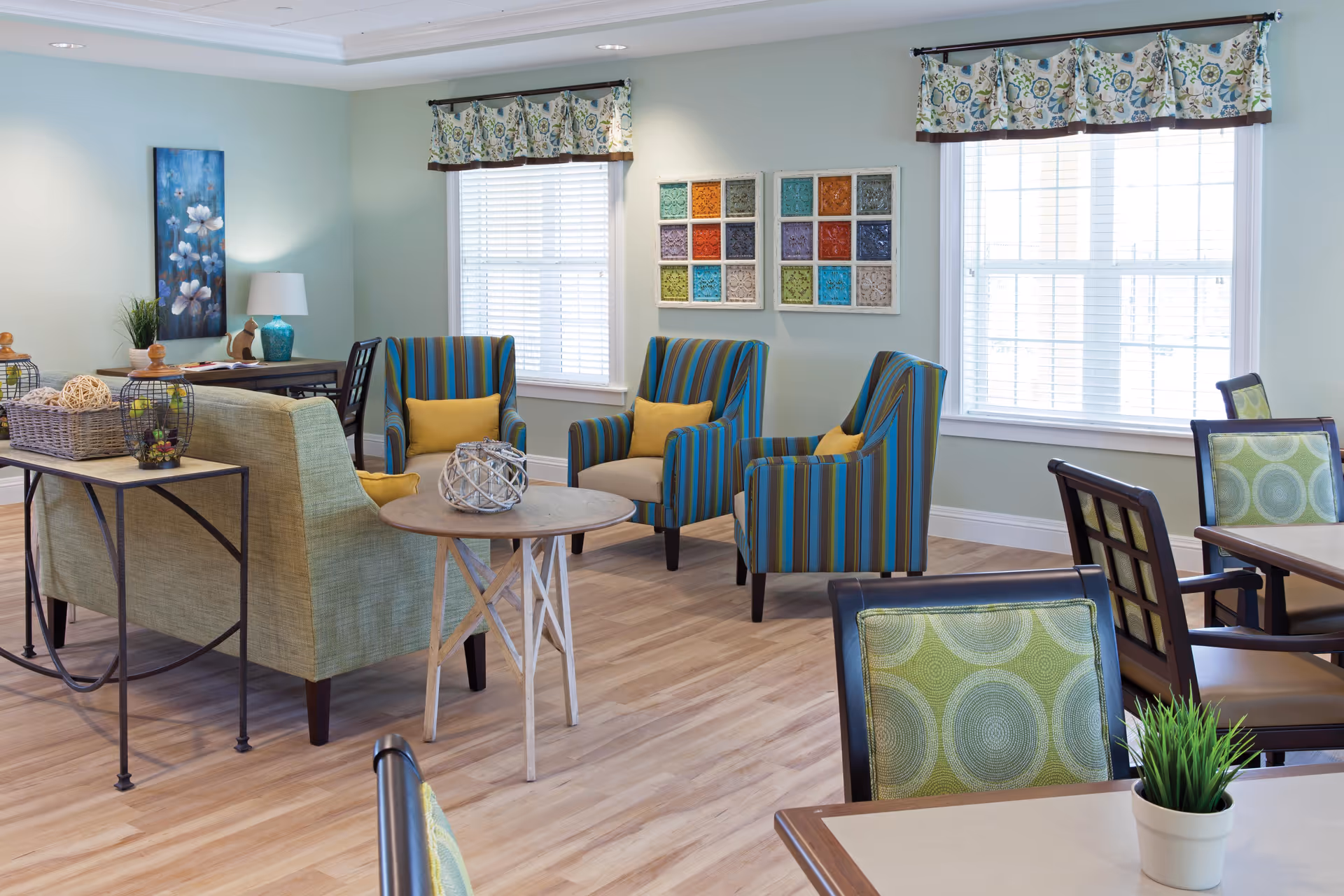 A bright and inviting common area with light wood flooring, featuring a green sofa, three striped armchairs with yellow cushions, and several dining tables with green patterned chairs. The room has two large windows with floral valances, colorful wall art, and decorative items on tables.
