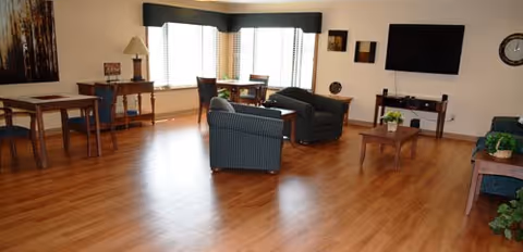 A spacious living room with wooden flooring featuring two striped armchairs, a coffee table with a small plant, a wall-mounted flat-screen TV, and several tables and chairs near large windows with blinds. The room is decorated with framed artwork, a clock, and various plants.