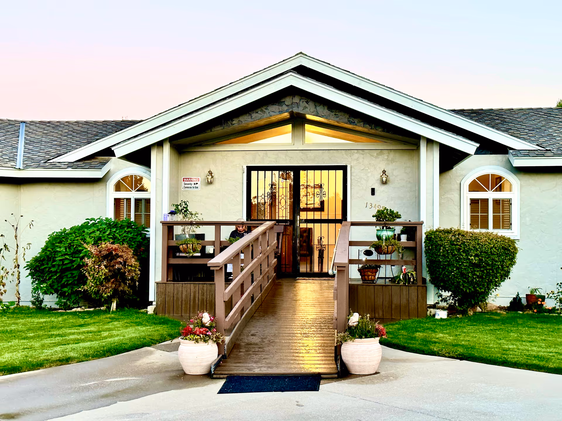 Front entrance of a residential care home with a wooden ramp leading to double doors with a security gate, flanked by potted plants and trimmed shrubs.