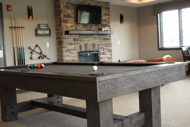 A recreational room featuring a dark wood pool table with billiard balls arranged on it. The room has a stone fireplace with a mounted TV above it, a rack holding pool cues on the wall, and a window with blinds letting in natural light. There are also some chairs and a small table near the window.