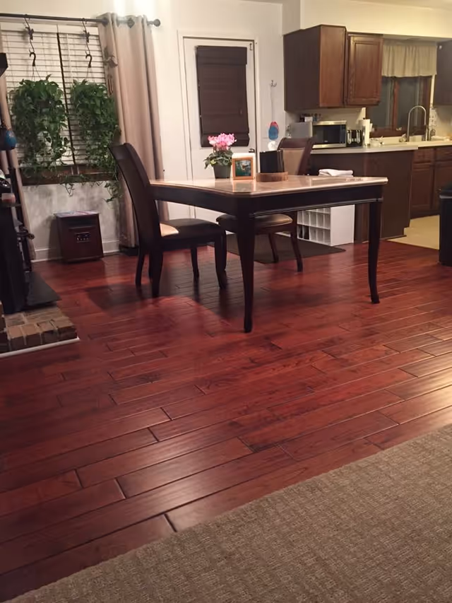 Interior view of a dining area with a wooden table and two chairs on a polished hardwood floor. A small potted plant and a framed photo are on the table. In the background, there is a kitchen with wooden cabinets, a microwave, and a sink. A window with blinds and curtains is visible, along with some hanging plants near the window.