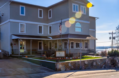 Two-story gray senior living facility with a porch, flagpole, and landscaped front garden.