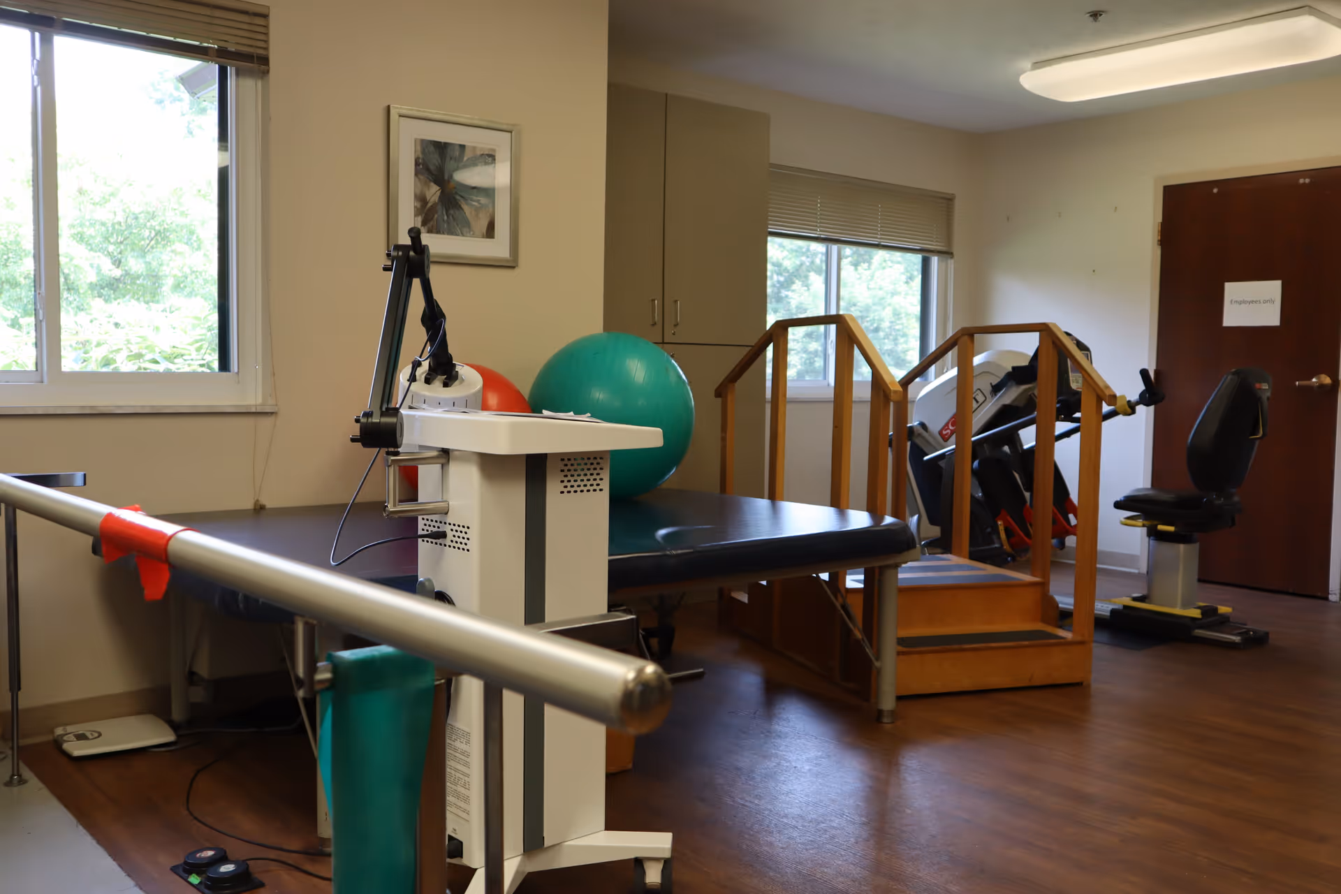 Physical therapy room with parallel bars, exercise balls, a treatment table, a small wooden staircase with handrails, and exercise equipment. The room has windows with blinds, wooden flooring, and a door labeled 'Employees only'.