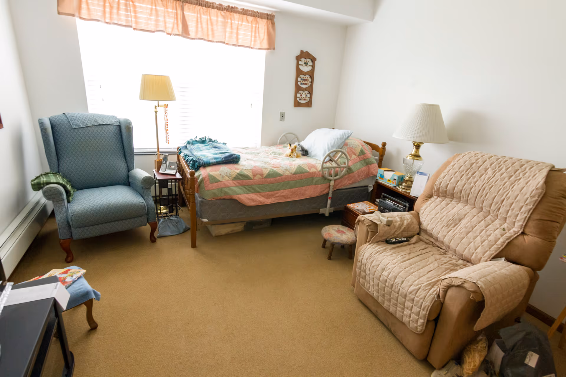 Small senior bedroom with a single bed covered by a quilt, two armchairs, lamps, and bedside personal items.