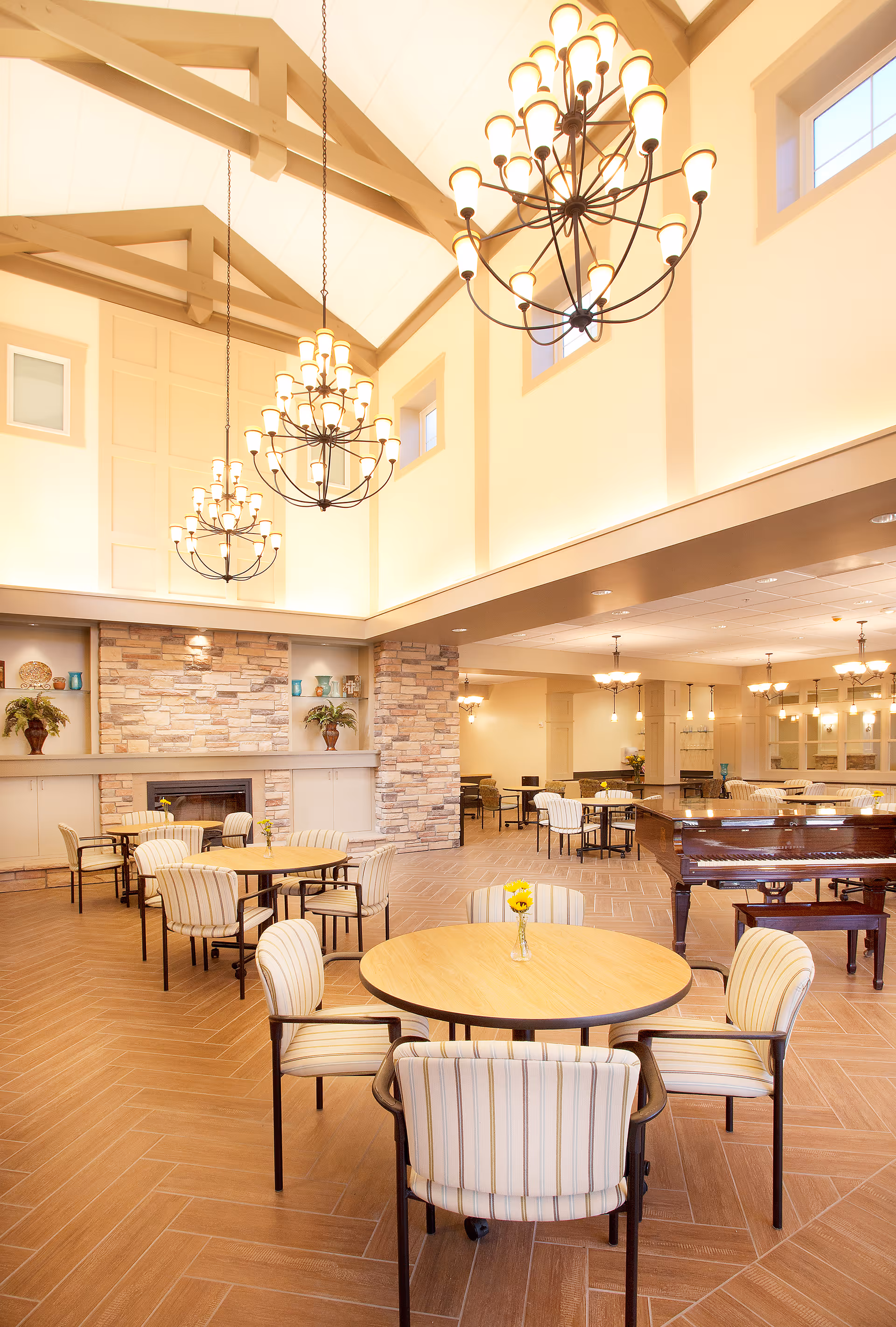 A spacious and well-lit common area with high vaulted ceilings and large chandeliers. The room features round tables with striped cushioned chairs arranged around them. There is a stone fireplace with decorative plants and shelves on either side. A grand piano is positioned to the right side of the room. The flooring is wood-patterned tile, and the walls are painted in warm neutral tones.