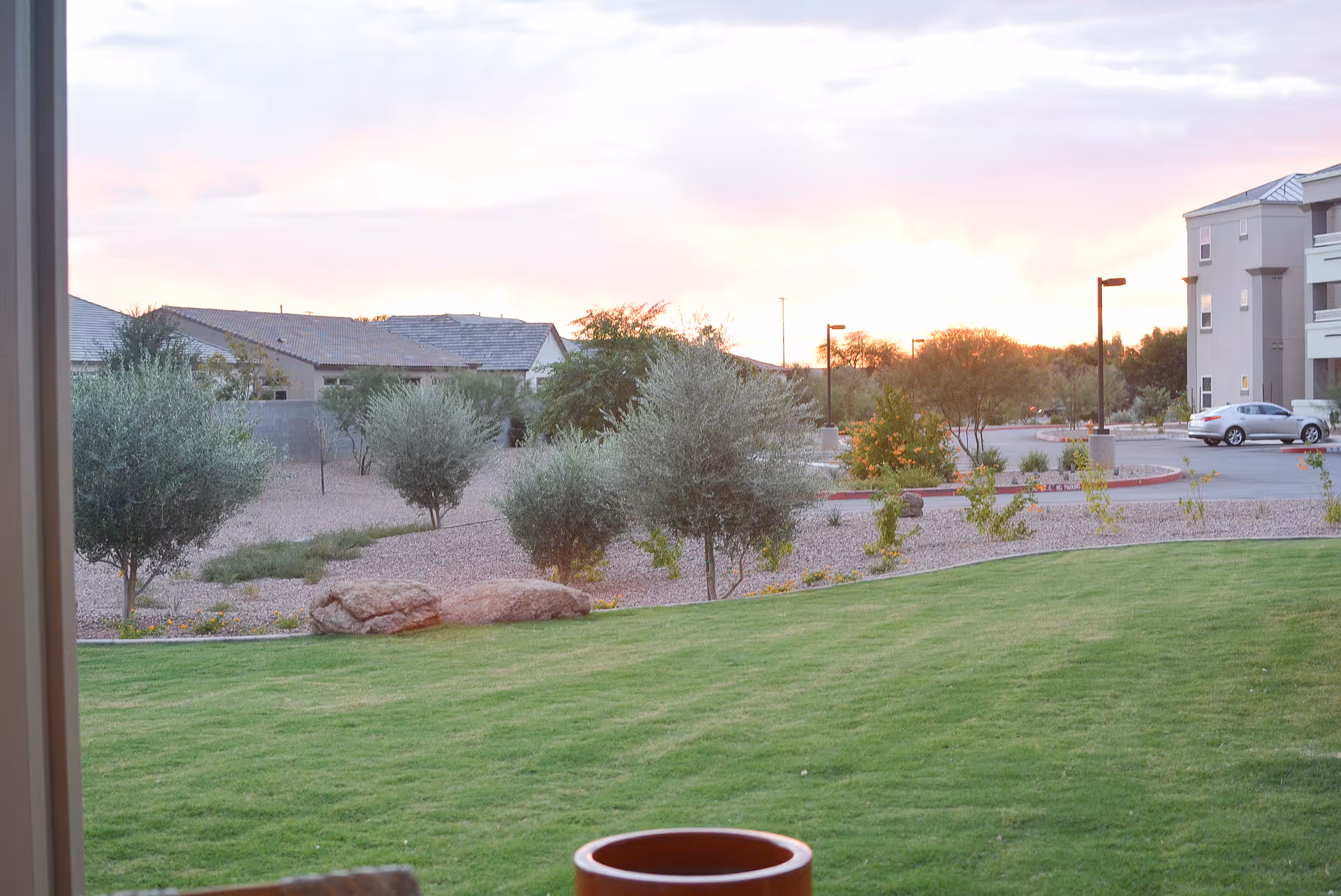 View of a landscaped outdoor area with green grass, small trees, and shrubs. In the background, there are residential buildings and a parking lot with a few cars. The sky is partly cloudy with a soft sunset glow.